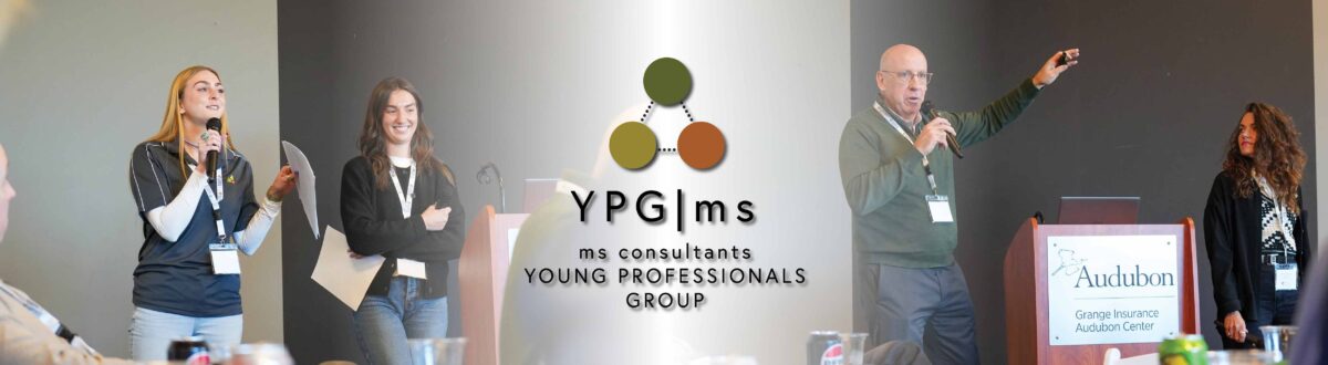 Two people standing at the front of a conference room speaking. A graphic sits in between the two people, explaining the young professionals group